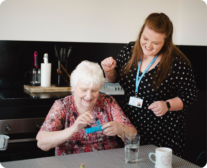 Caregiver supporting an older woman as she opens her pill organiser at home