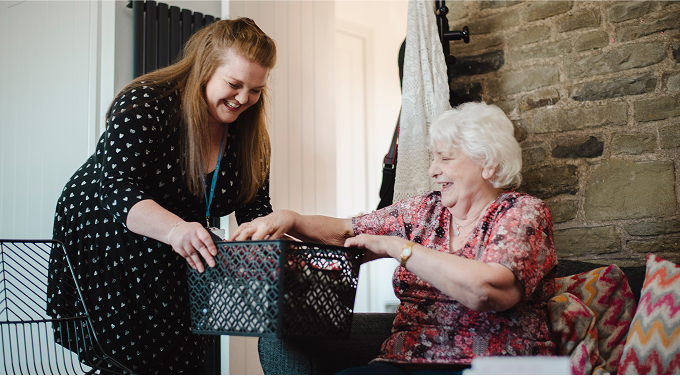 smiling carer passing box to elderly woman sat on sofa
